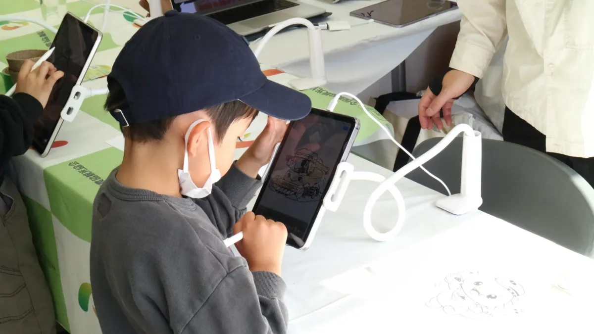 マスクとキャップを着用した子供が、スタイラスを使ってタブレットに絵を描いている様子