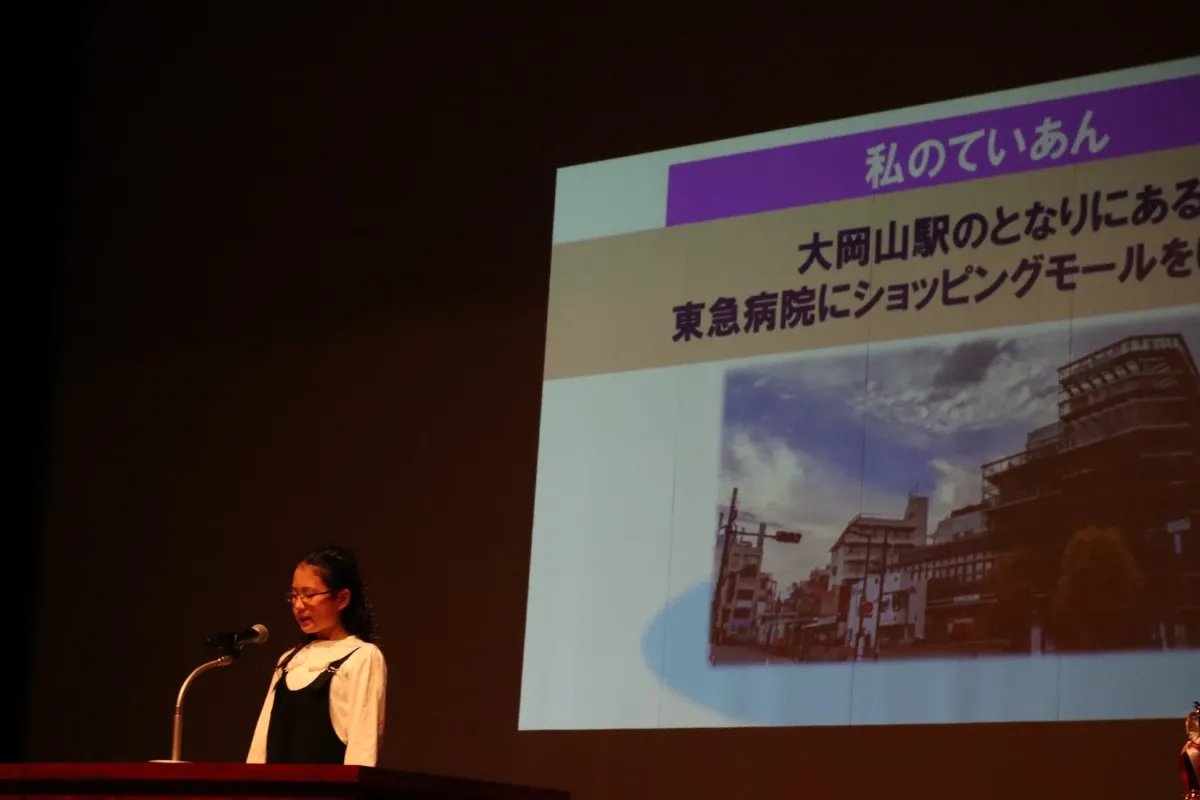 ステージ上で発表者がプロジェクター画面を背景にプレゼンテーションを行っています。画面には「私のていあん」「大岡山駅のとなりにある」「東急病院にショッピングモールを」というテキストと、街並みの写真が表示されています。