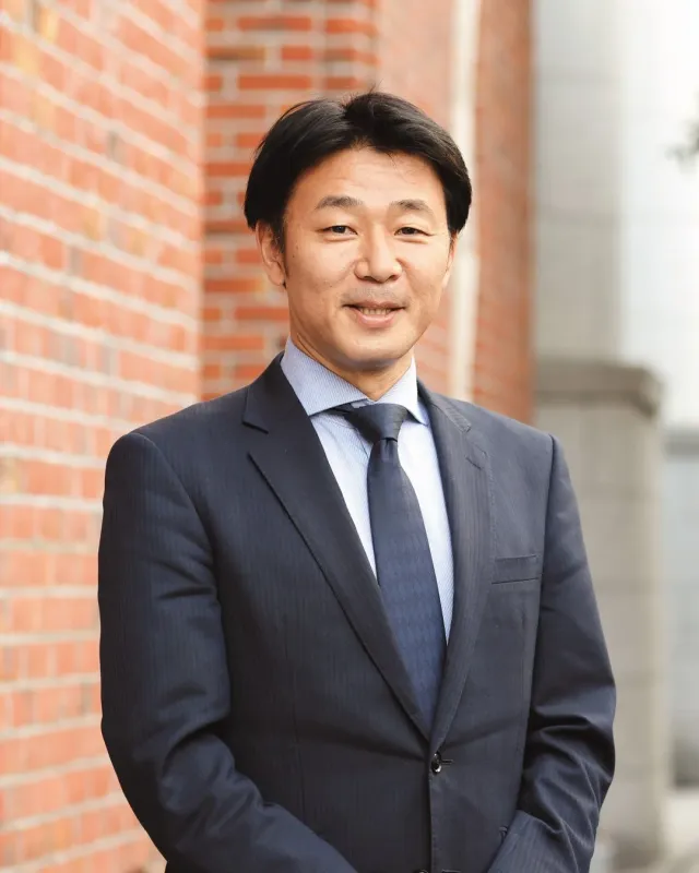 ダークスーツと水色のシャツを着た男性が、レンガの壁を背景に屋外でカメラに向かって微笑んでいます。