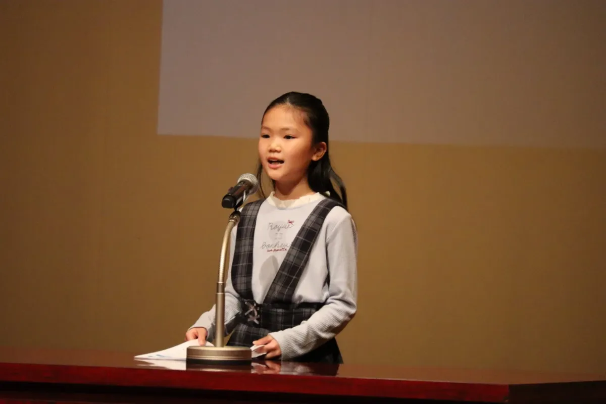 マイクの前に立つ少女がスピーチをしている様子です。壇上で書類を手に持ち、真剣な表情で話しています。学校の発表会やイベントでの一幕と思われます。