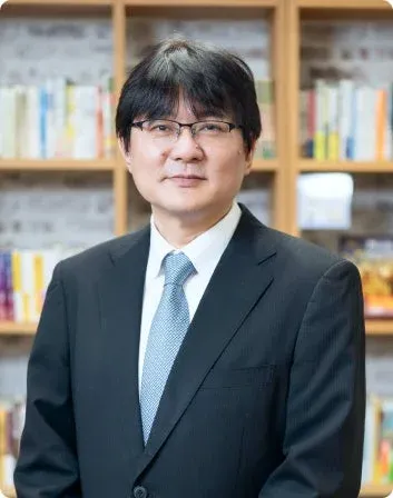 眼鏡をかけた中年の男性がスーツ姿で書棚を背景に立つポートレート