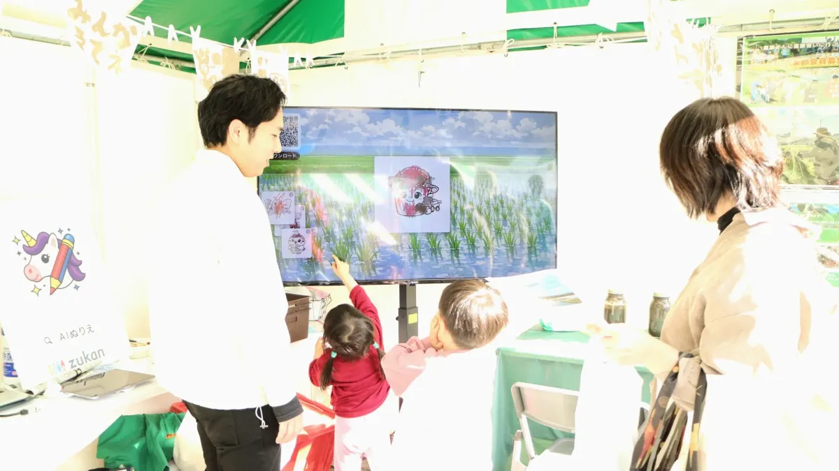 イベント会場のブースで、男性と子供たちがAIぬりえが体験できる大型ディスプレイを見ている様子