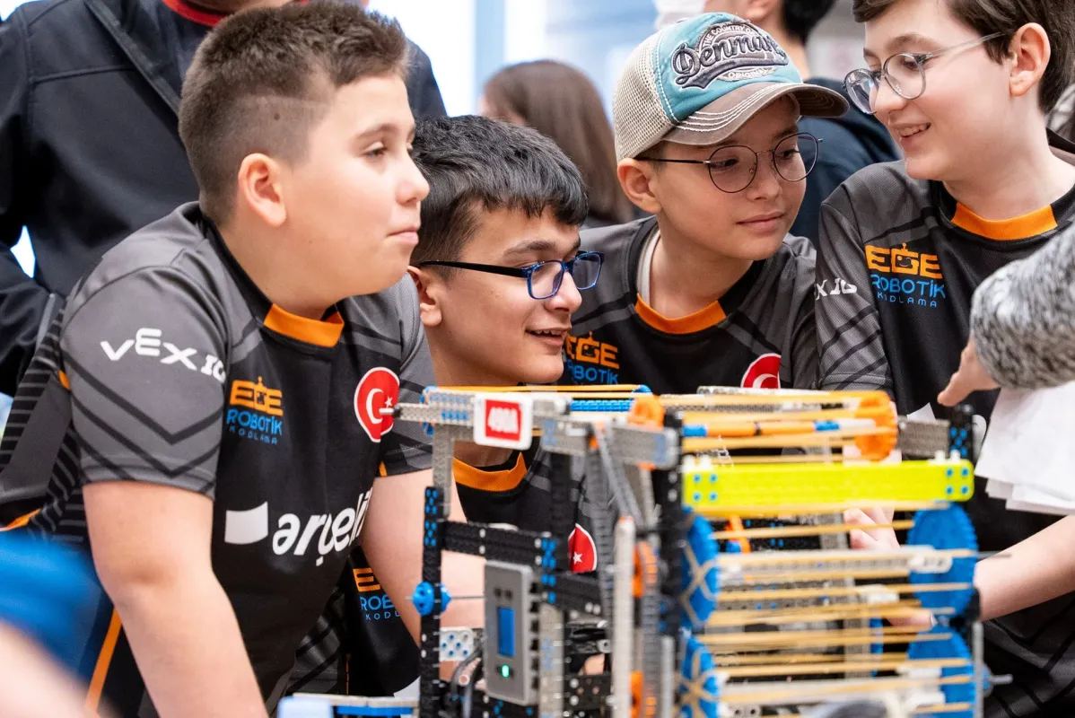 VEX IQとEGE ROBOTIKのロゴ入りシャツを着た少年たちが、ロボット競技会で複雑なロボットに集中し、協力し合っている様子