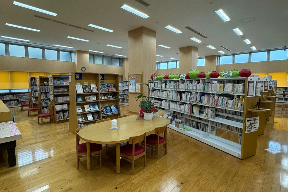 広々とした図書館の内部で、多くの本棚が並び、中央には読書用の大きな木製テーブルと椅子が配置されています。