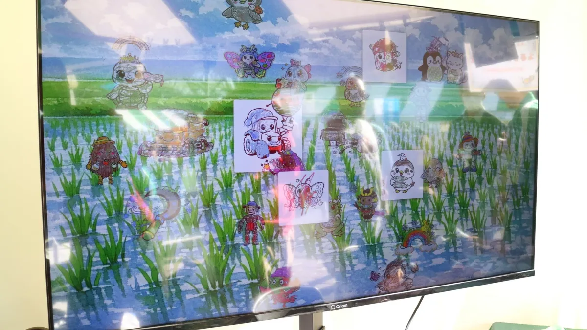 水田の背景が映るテレビ画面に、子供が描いたような可愛らしいキャラクターイラストが多数配置されています