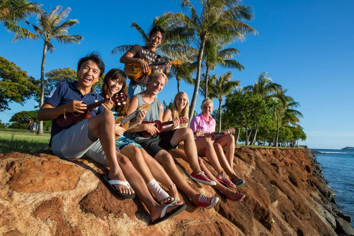 熱帯の青空の下、ヤシの木が並ぶ海岸で、6人の若者たちがウクレレを演奏しながら楽しそうに笑っています。