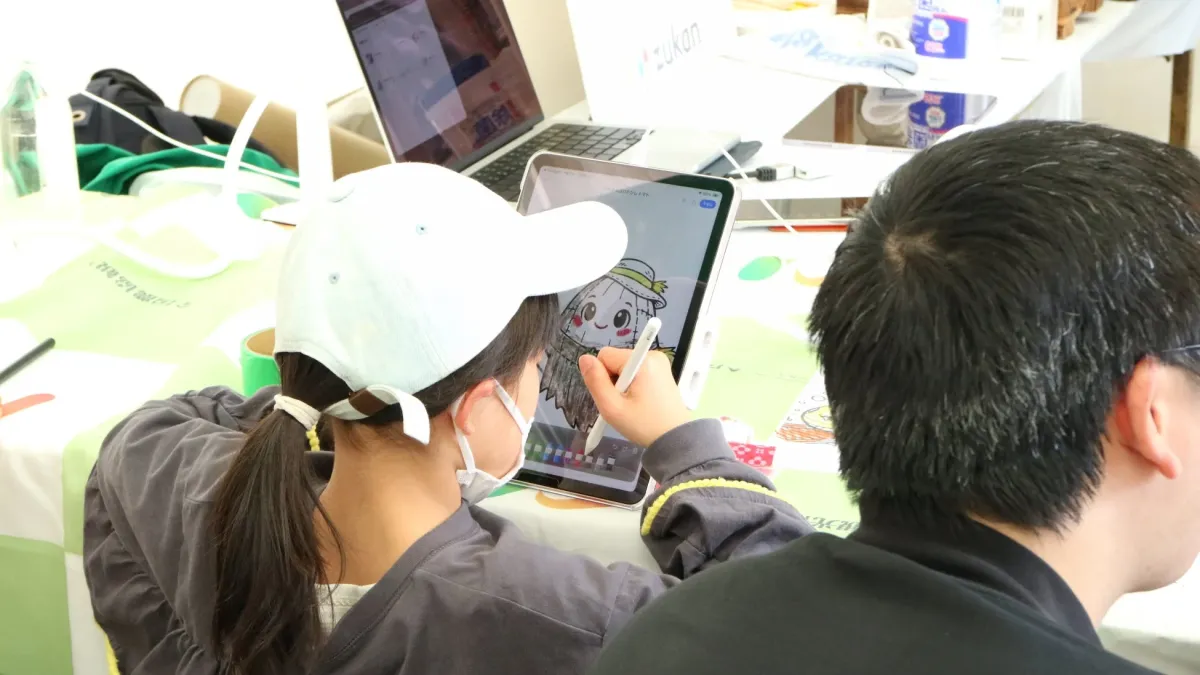 キャップとマスクを着用した子供が、イベント会場のような場所でiPadとApple Pencilを使ってかかしのイラストを熱心に描いています