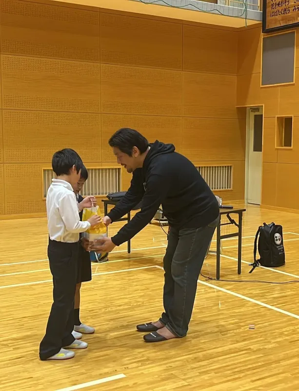 体育館のような屋内空間で、大人の男性が二人の子供に黄色い袋に入った物を手渡しています。