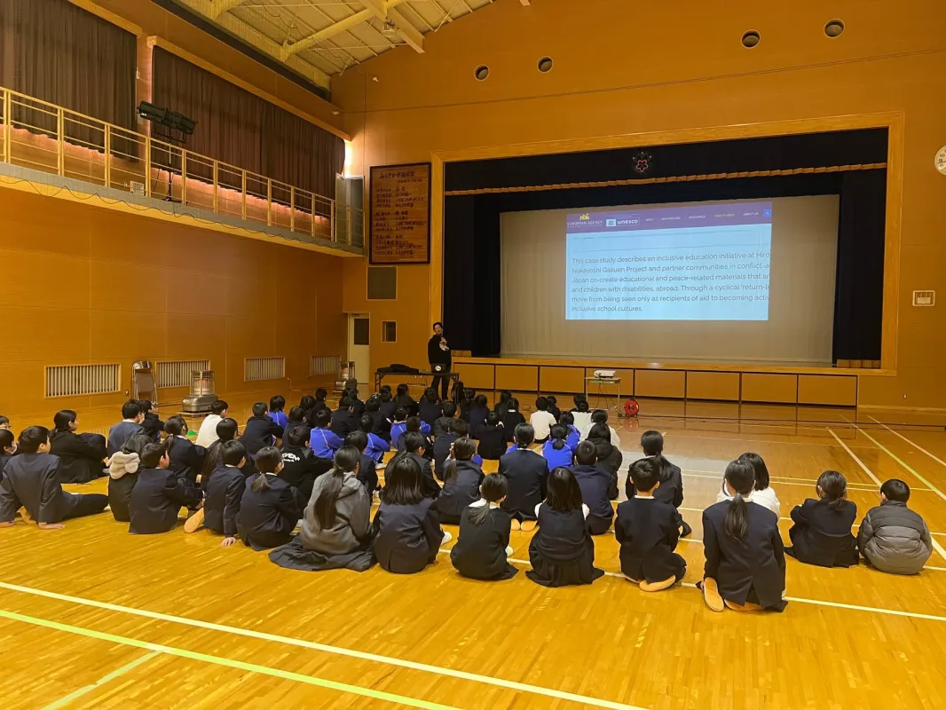 体育館で多くの生徒が床に座り、壇上の講師によるプレゼンテーションを聞いている